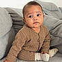 baby, child, infant, sitting, brown_clothing, ribbed_sweater, grey_blanket, grey_cushions, indoor, portrait, cute, young_child, cozy, soft_texture, hand, face, expression, looking_away, sock, floor