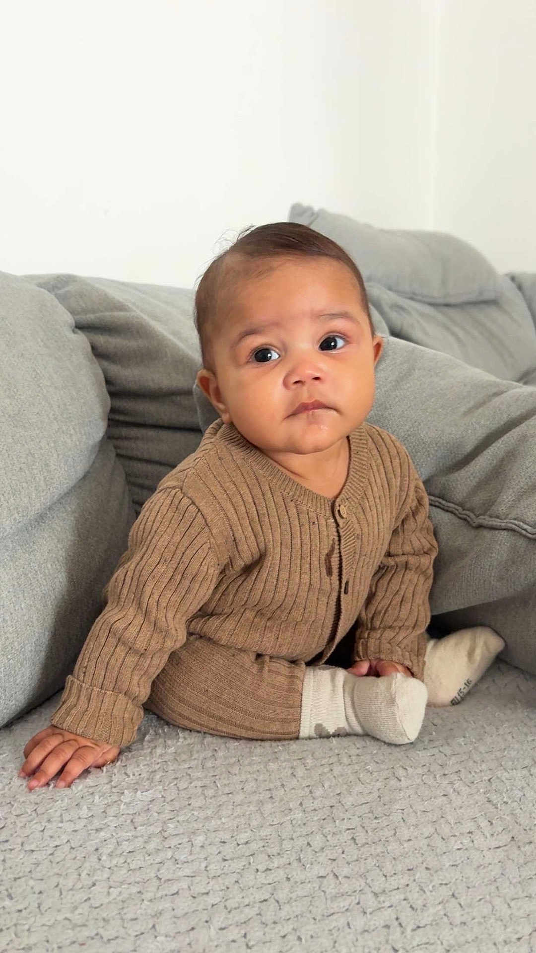 Anastasya a rejoint le concours — aidez-le/la à gagner de superbes lots ! baby, child, infant, sitting, brown_clothing, ribbed_sweater, grey_blanket, grey_cushions, indoor, portrait, cute, young_child, cozy, soft_texture, hand, face, expression, looking_away, sock, floor