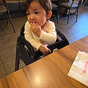 Eliana joined the competition — help win amazing prizes! black_chair, casual, chair, child, curly_hair, eating, floor, hand_in_mouth, high_chair, indoor, long_sleeves, person, restaurant, seated, table, tile_floor, toddler, white_clothing, wooden_table, young_child