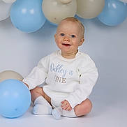 Oakley is registered to the contest to win money with this photo: baby, balloon, beige_balloon, blue_balloon, celebration, child, cute, decorations, happy, infant, one_year_old, onesie, party, portrait, sitting, smiling, sock, soft_lighting, studio, white_clothing