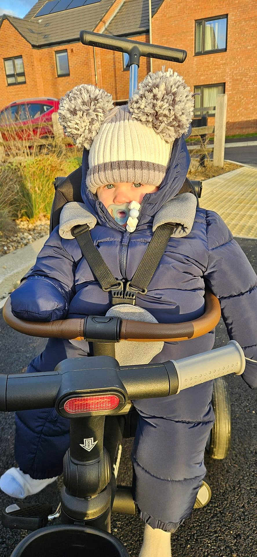 Oakley is registered to the contest to win money with this photo: toddler, child, hat, pom_poms, pacifier, snowsuit, jacket, stroller, tricycle, outdoor, sunlight, sidewalk, residential_area, toy, winter_clothing, baby, seatbelt, wheel, road, cute