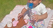 Octavia is registered to the contest to win money with this photo: toddler, child, pacifier, curly_hair, outdoor, blanket, stuffed_animal, toy, playtime, grass, sunlight, happy, baby_clothes, floral_pants, plush, smiling, barefoot, nature, garden, daytime