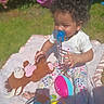 toddler, child, pacifier, curly_hair, outdoor, blanket, stuffed_animal, toy, playtime, grass, sunlight, happy, baby_clothes, floral_pants, plush, smiling, barefoot, nature, garden, daytime