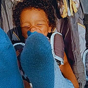 Octavia is registered to the contest to win money with this photo: child, curly_hair, smiling, blue_sock, feet, stroller, seatbelt, happy, infant, baby, clothing, pattern, fabric, seat, playful, cute, portrait, outdoor, sunlight, tan_skin
