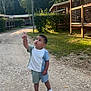 cabin, child, curious, daytime, gravel, greenery, nature, outdoor, park, path, person, pointing, shorts, sneakers, summer, sunlight, toddler, trees, tshirt, wooden_structure