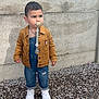 casual, child, clothing, concrete_wall, denim_overalls, fashion, footwear, ground, jacket, outdoor, pacifier, pebbles, person, serious_expression, standing, texture, toddler, wear, white_sneakers, young_child