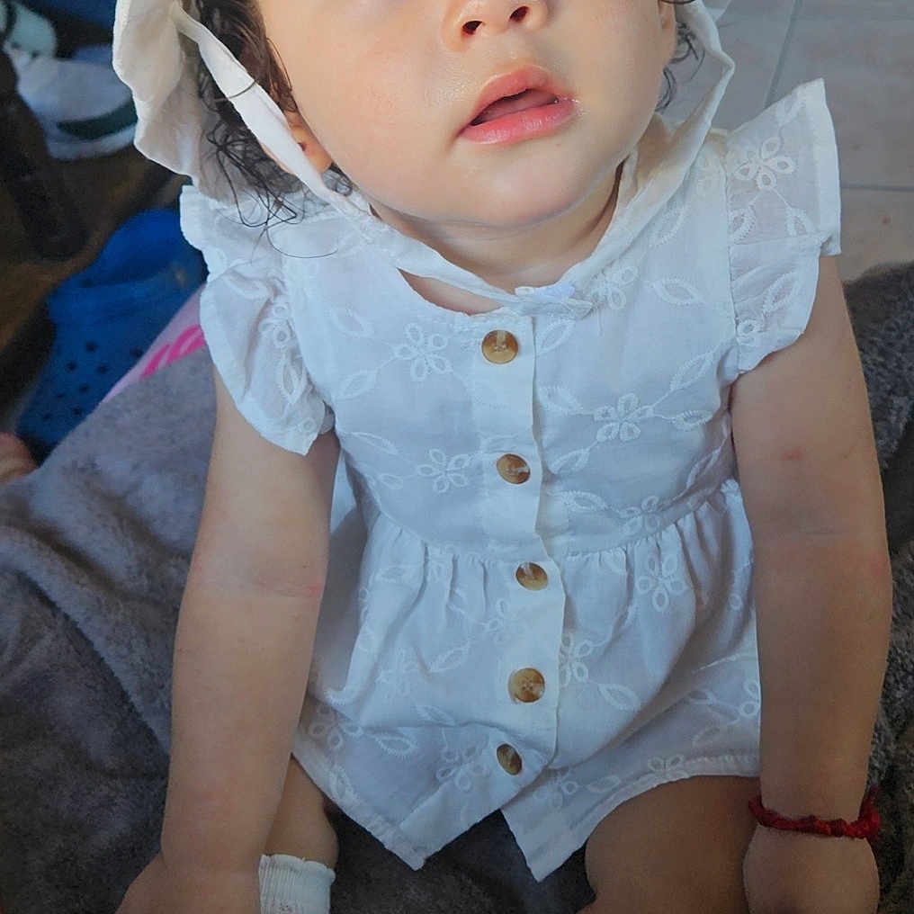 Daylene is registered to the contest to win money with this photo: baby, blanket, bonnet, buttoned_dress, casual, child, cozy, curly_hair, cute, expression, floor, home, indoors, person, portrait, sitting, skin, sock, toddler, white_dress