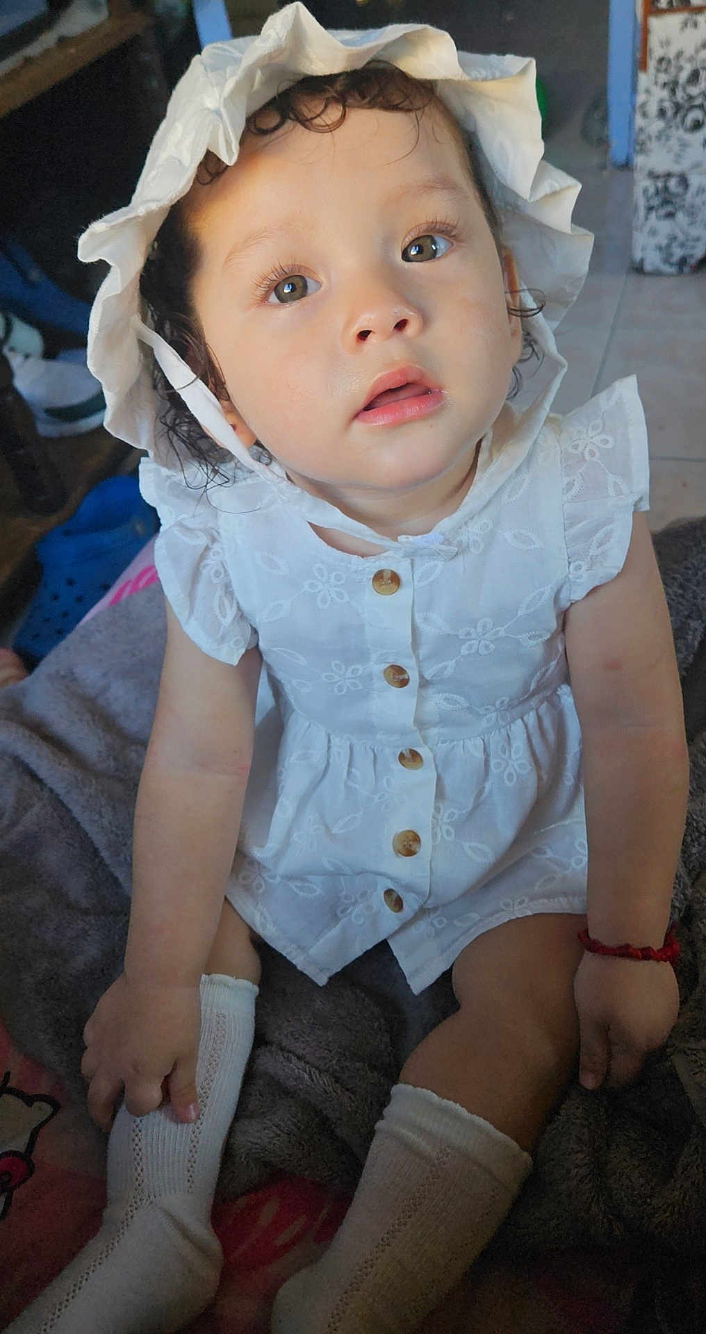 Daylene is registered to the contest to win money with this photo: baby, blanket, bonnet, buttoned_dress, casual, child, cozy, curly_hair, cute, expression, floor, home, indoors, person, portrait, sitting, skin, sock, toddler, white_dress