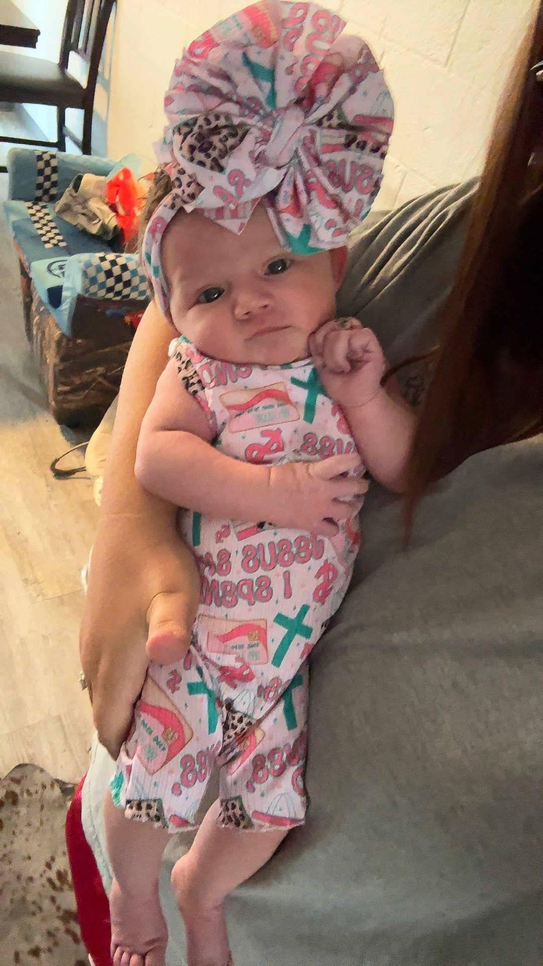 Madison joined the competition — help win amazing prizes! baby, infant, child, person, holding, headband, bow, clothing, patterned_outfit, indoor, adult_arm, floor, carpet, furniture, couch, wooden_chair, curious, portrait, closeup, home