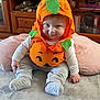 baby, child, pumpkin_costume, orange, green, smile, cute, sitting, cushion, soft_surface, indoors, clothing, hood, face, expression, playful, person, infant, home, cozy