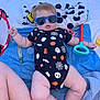 baby, sunglasses, onesie, halloween, toy_shovel, towel, outdoor, child, infant, legs, arm, plastic_toy, blue, black_clothing, white_towel, patterned_clothing, sunlight, relaxing, cute, playtime