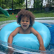Jamari joined the competition — help win amazing prizes! child, pool, inflatable_ring, water, curly_hair, smiling, outdoor, summer, backyard, slide, grass, trees, fence, splashing, happy, play, fun, person, daytime, recreation