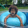 child, pool, inflatable_ring, water, curly_hair, smiling, outdoor, summer, backyard, slide, grass, trees, fence, splashing, happy, play, fun, person, daytime, recreation