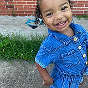 Kailani joined the competition — help win amazing prizes! child, toddler, denim_dress, smile, curly_hair, outdoor, grass, concrete, brick_wall, happy, person, cute, portrait, playful, young_child, fashion, feet, buttoned_clothing, standing, sunlight