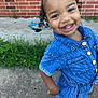child, toddler, denim_dress, smile, curly_hair, outdoor, grass, concrete, brick_wall, happy, person, cute, portrait, playful, young_child, fashion, feet, buttoned_clothing, standing, sunlight