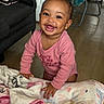 baby, smile, pink_onesie, blanket, cushion, floor, happy, indoor, child, person, earring, wood_floor, furniture, home, cute, portrait, toddler, expression, cozy, infant