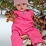 Alannah Carol is registered to the contest to win money with this photo: baby, child, cold_weather, curious_expression, cute, face, greenery, hand, holding, infant, knitted_hat, leaves, outdoor, person, pink_overalls, pom_pom, portrait, snow, socks, winter_clothing