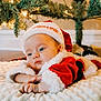 Jaxson is registered to the contest to win money with this photo: baby, christmas, santa_hat, red_clothing, blanket, infant, holiday, decorations, tree, lights, cute, indoors, soft_texture, face, hands, festive, warm, cozy, child, portrait