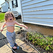 Holden is registered to the contest to win money with this photo: barefoot, child, concrete, curly_hair, daytime, garden, greenery, happy, hose, nature, outdoor, pants, plants, playful, side_of_house, smiling, succulents, sunny, toddler, water