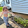 barefoot, child, concrete, curly_hair, daytime, garden, greenery, happy, hose, nature, outdoor, pants, plants, playful, side_of_house, smiling, succulents, sunny, toddler, water
