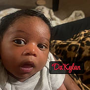 Dakylan joined the competition — help win amazing prizes! baby, infant, child, portrait, close_up, face, eyes, looking_at_camera, curious, indoor, bedroom, sitting, being_held, adult_arm, white_onesie, blanket, leopard_print, bedding, name_text, photograph