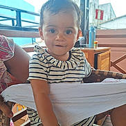 Alice a rejoint le concours — aidez-le/la à gagner de superbes lots ! toddler, child, striped_clothing, curly_hair, indoor, chair, wooden_furniture, hand, footwear, person, baby, smiling, cute, young_child, casual_wear, daylight, portrait, expression, comfortable, curious