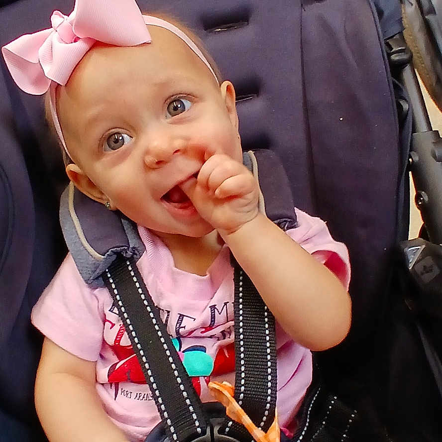 Thyra Eddy Dawn Loza is registered to the contest to win money with this photo: baby, child, closeup, cute, face, happy, harness, headband, indoor, infant, person, pink_bow, pink_shirt, playful, seated, small_child, smiling, stroller, sucking_thumb, toy