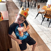 Eli is registered to the contest to win money with this photo: boy, burger_king, casual_clothing, chairs, child, crown, cup, dining_area, drink, fast_food, indoors, paper_crown, portrait, restaurant, serious_expression, sitting, standing, straw, tables, tiled_floor