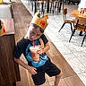 boy, burger_king, casual_clothing, chairs, child, crown, cup, dining_area, drink, fast_food, indoors, paper_crown, portrait, restaurant, serious_expression, sitting, standing, straw, tables, tiled_floor
