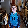 child, boy, young, smiling, portrait, indoors, kitchen, hoodie, blue_clothing, standing, happy, face, brown_hair, eyes, refrigerator, dining_chair, wooden_floor, trash_bin, stroller, home