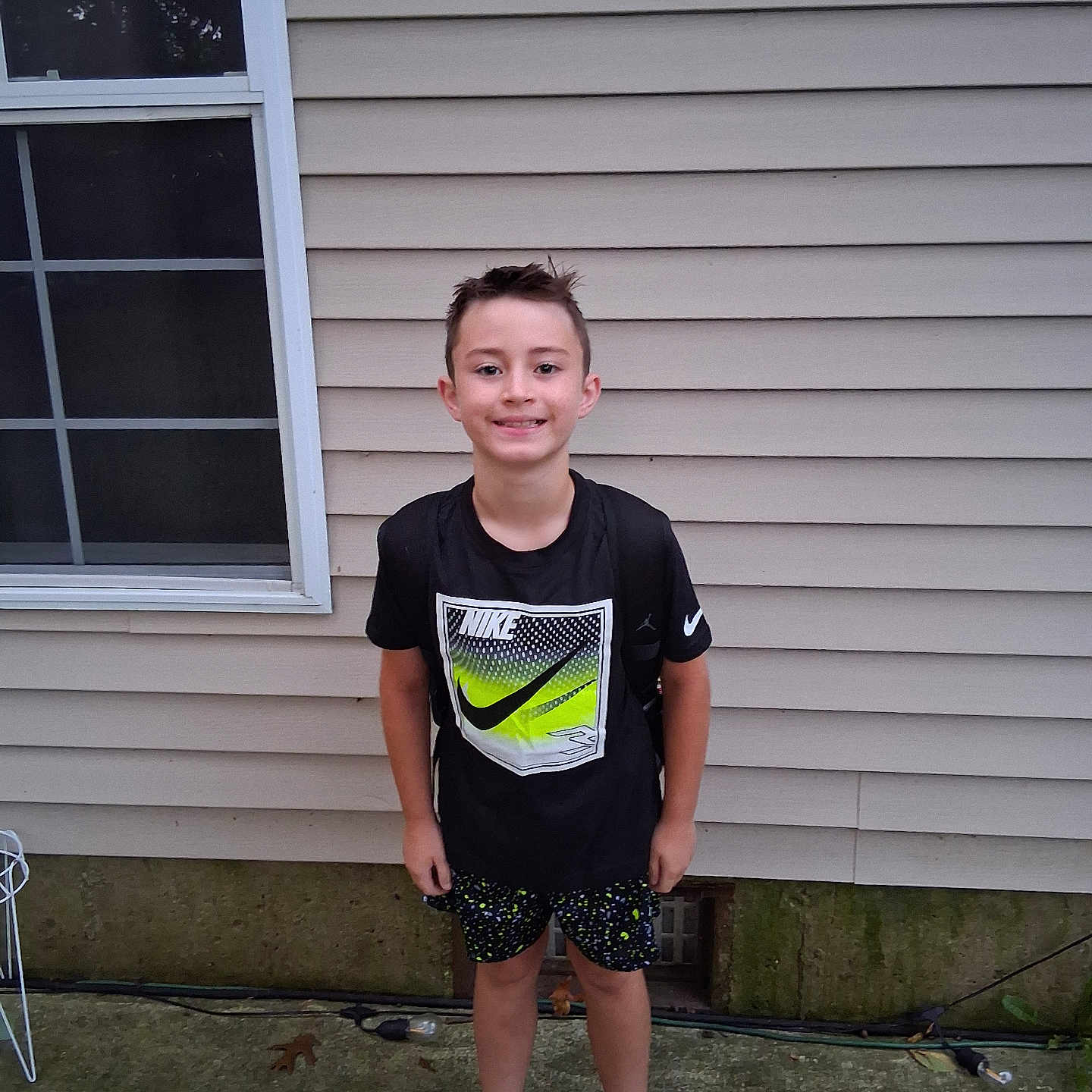Eli is registered to the contest to win money with this photo: child, boy, smiling, portrait, outdoor, standing, house, siding, window, patio, concrete, sneakers, shoes, sportswear, t_shirt, shorts, nike, suburban, daylight, solo