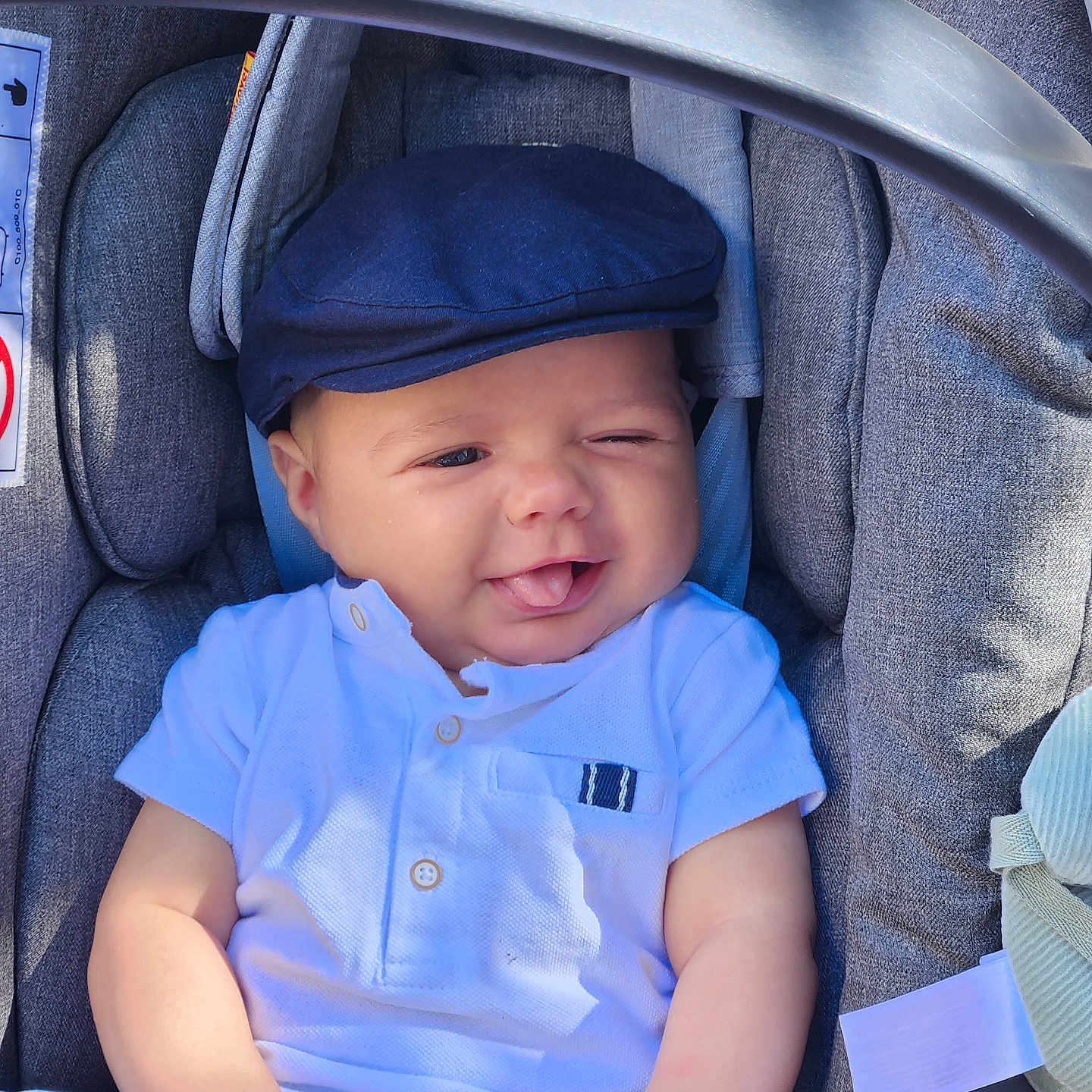 Jules a rejoint le concours — aidez-le/la à gagner de superbes lots ! baby, blue_shorts, cap, car_seat, child, clothing, comfort, cute, fabric, indoors, infant, person, playful, portrait, seat_belt, smiling, tongue_out, white_shirt, winking, young_child