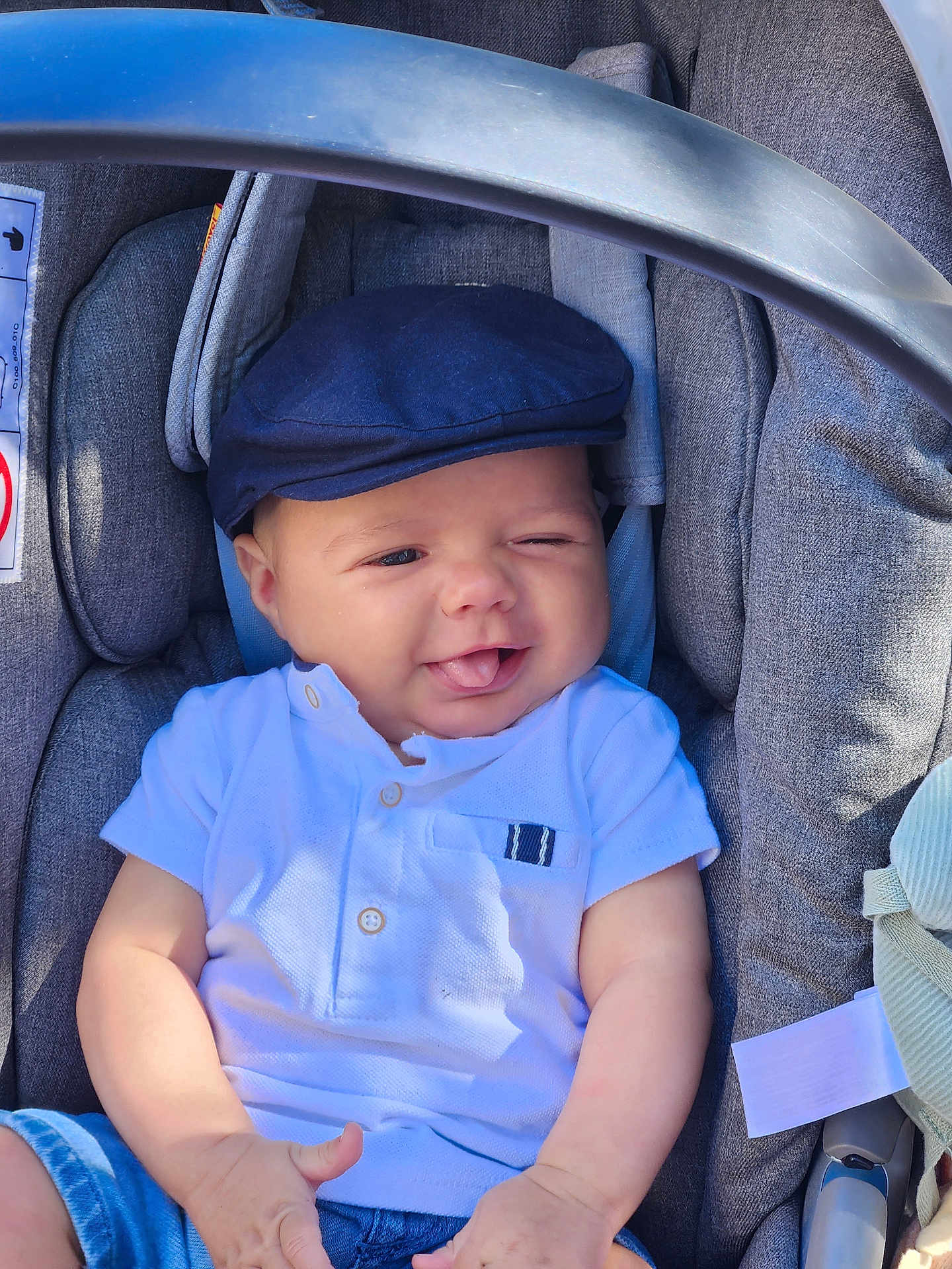 Jules a rejoint le concours — aidez-le/la à gagner de superbes lots ! baby, child, car_seat, cap, white_shirt, blue_shorts, tongue_out, winking, smiling, cute, infant, seat_belt, fabric, clothing, indoors, comfort, playful, portrait, person, young_child