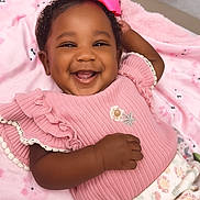 Ocean-Sinclair is registered to the contest to win money with this photo: baby, smiling, pink_bow, pink_clothing, blanket, happy, cute, infant, portrait, child, face, hand, curly_hair, cozy, soft_texture, lying_down, indoors, cheerful, ruffled_sleeves, patterned_pants