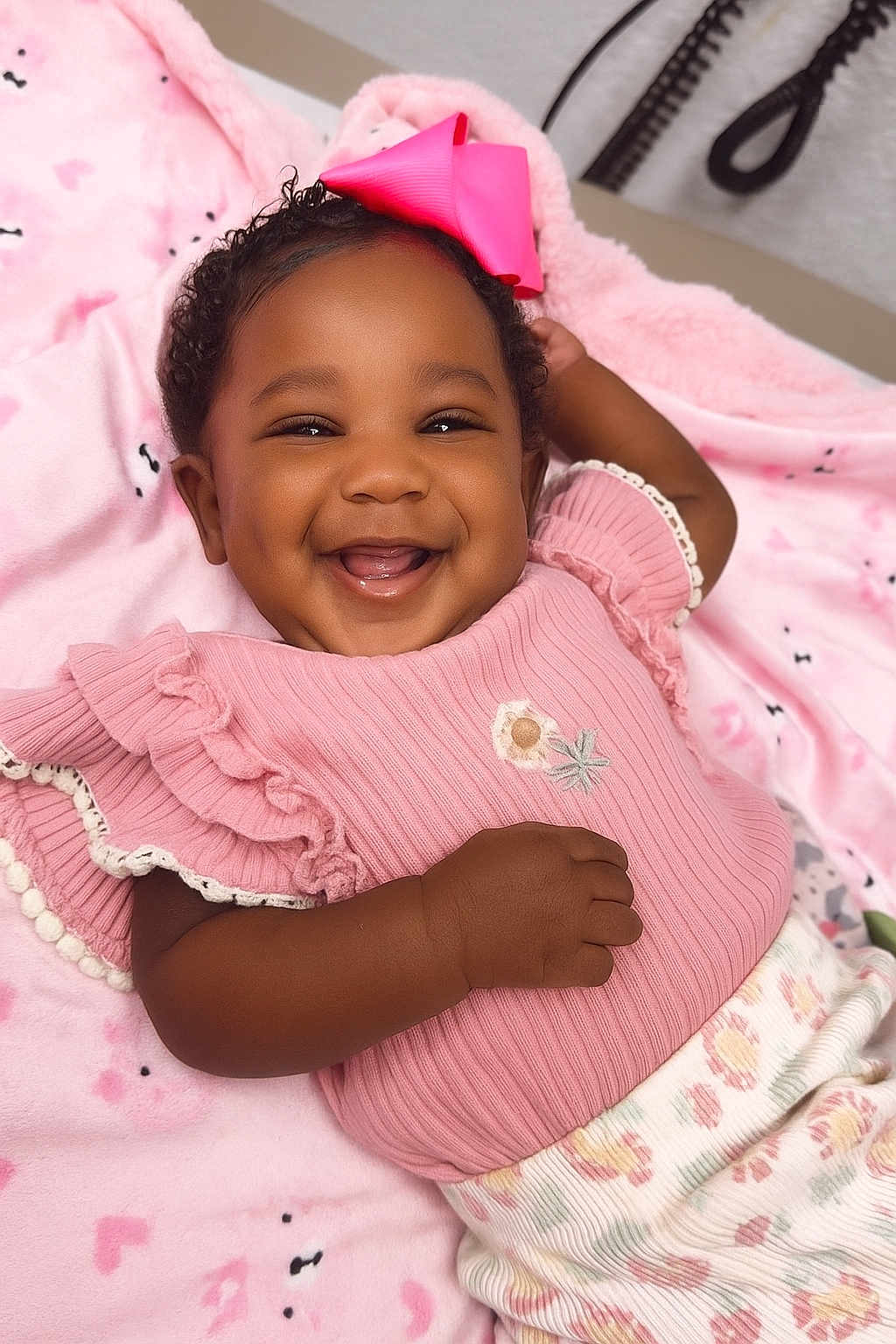 Ocean-Sinclair is registered to the contest to win money with this photo: baby, smiling, pink_bow, pink_clothing, blanket, happy, cute, infant, portrait, child, face, hand, curly_hair, cozy, soft_texture, lying_down, indoors, cheerful, ruffled_sleeves, patterned_pants
