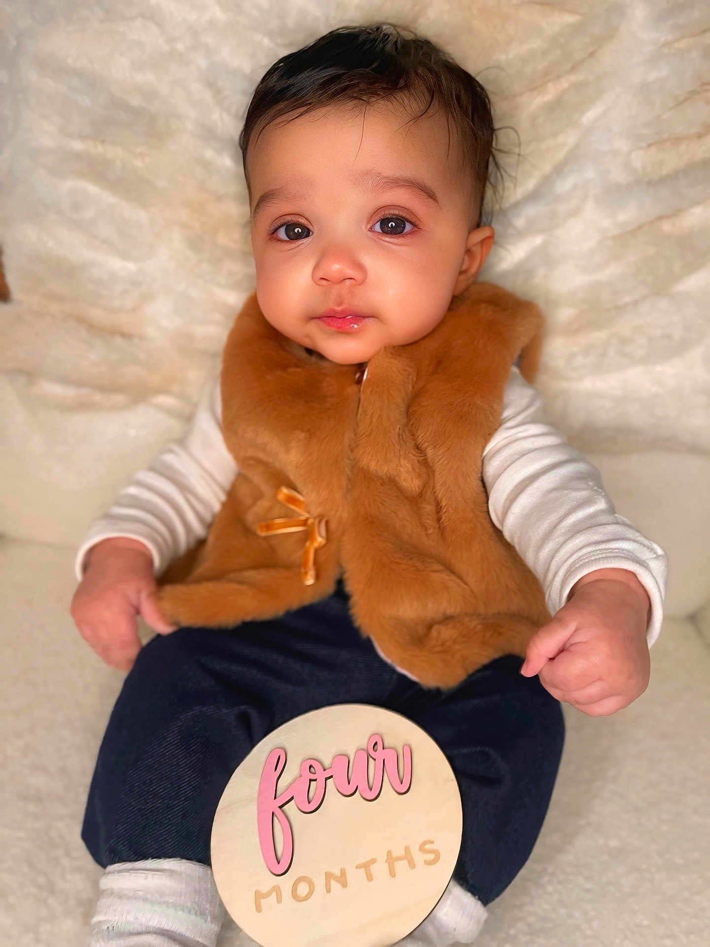 Gianna is registered to the contest to win money with this photo: baby, infant, child, four_months, milestone_sign, wooden_sign, fur_vest, brown_vest, blanket, portrait, indoor, cute, big_eyes, chubby_cheeks, sitting, hands, socks, dark_pants, soft_lighting, close_up