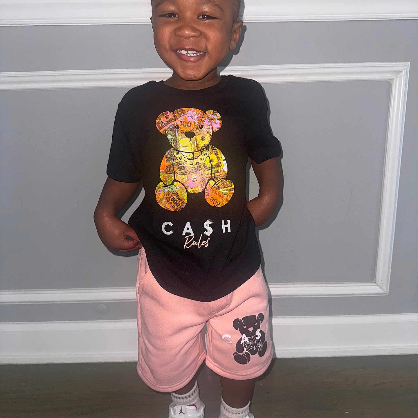Zyheir is registered to the contest to win money with this photo: black_tshirt, casual_clothing, child, cute, fashion, floor, footwear, happy, indoor, light_skin, person, pink_shorts, playful, portrait, short_hair, smiling, standing, toddler, wall, white_sneakers