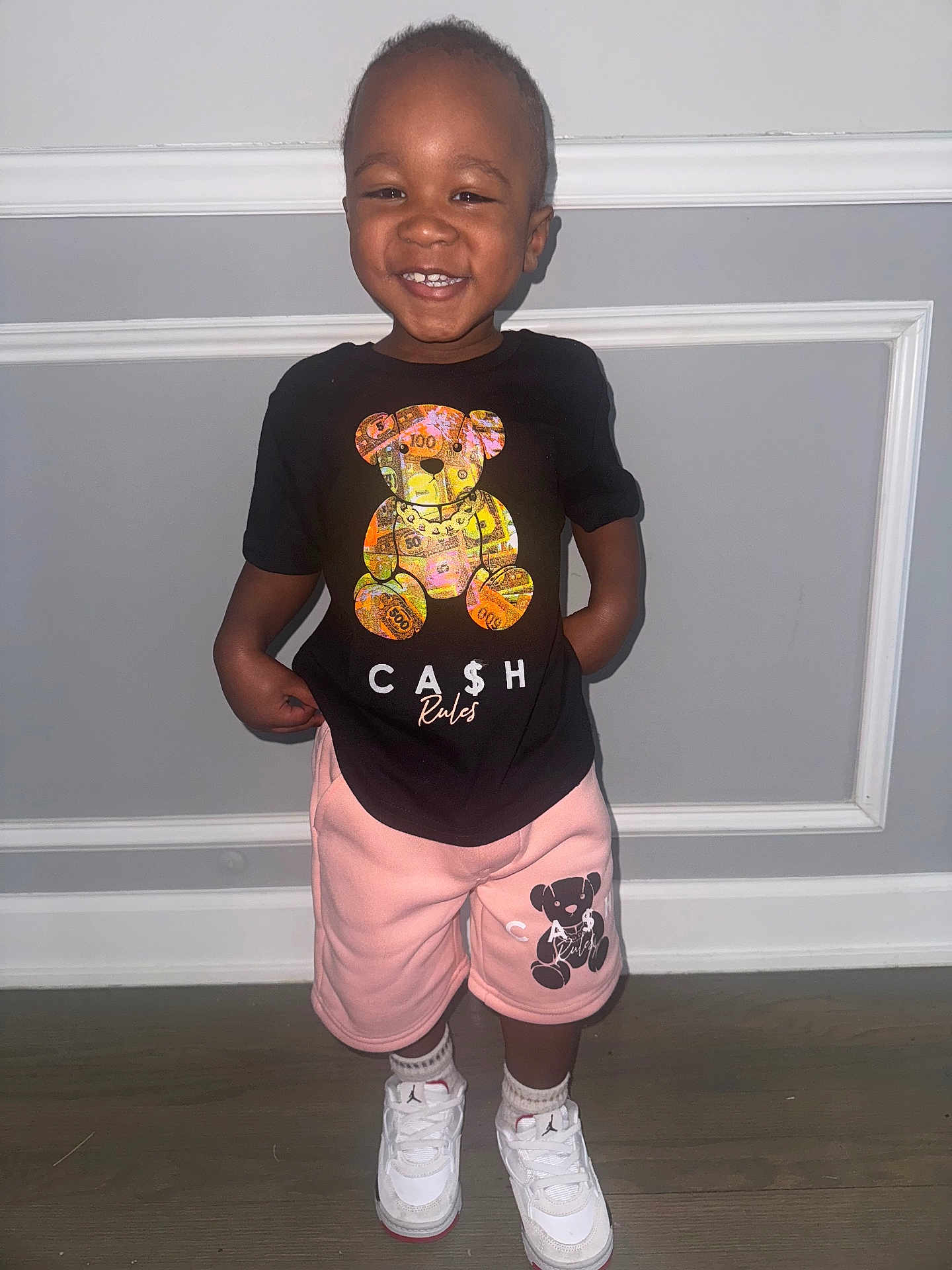 Zyheir is registered to the contest to win money with this photo: black_tshirt, casual_clothing, child, cute, fashion, floor, footwear, happy, indoor, light_skin, person, pink_shorts, playful, portrait, short_hair, smiling, standing, toddler, wall, white_sneakers