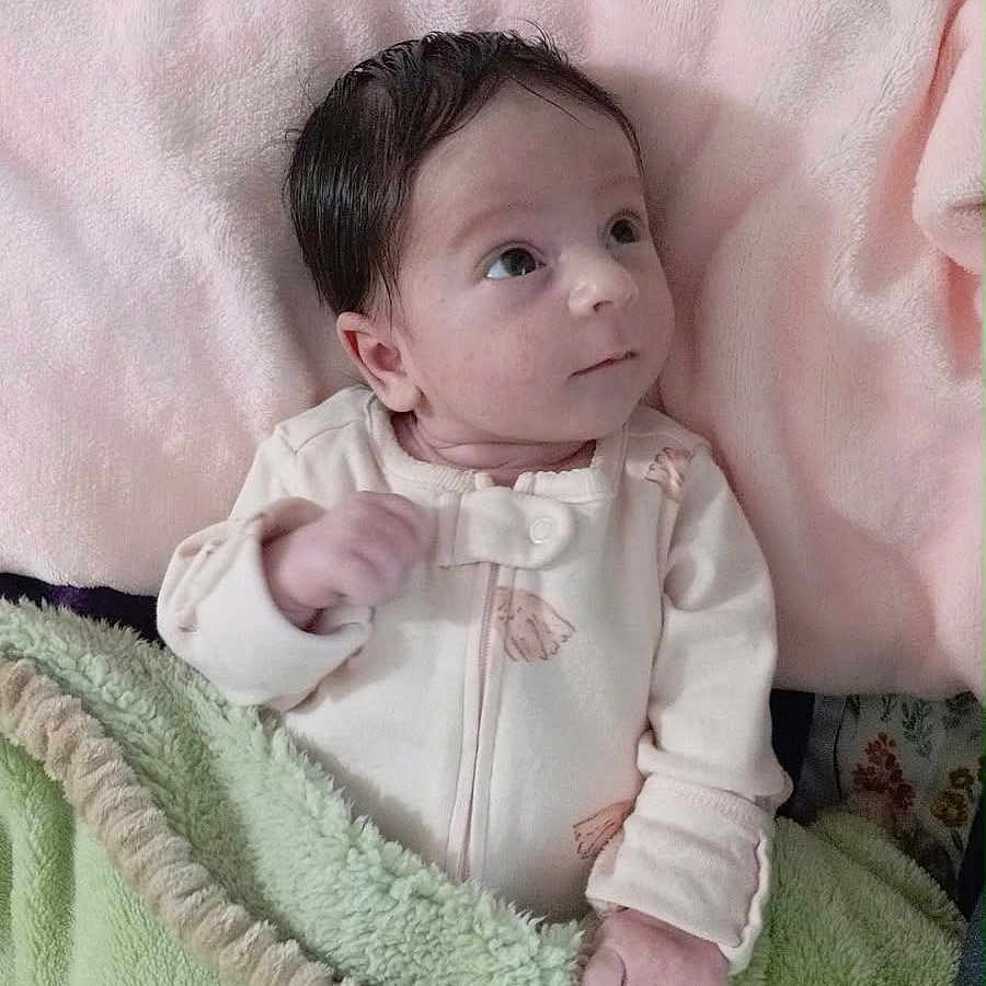Ausrine is registered to the contest to win money with this photo: baby, infant, child, portrait, indoors, nursery, blanket, pink_blanket, green_blanket, floral_background, baby_clothing, onesie, sitting, lying_down, wide_eyes, dark_hair, cozy, soft_furnishings, natural_light, close_up