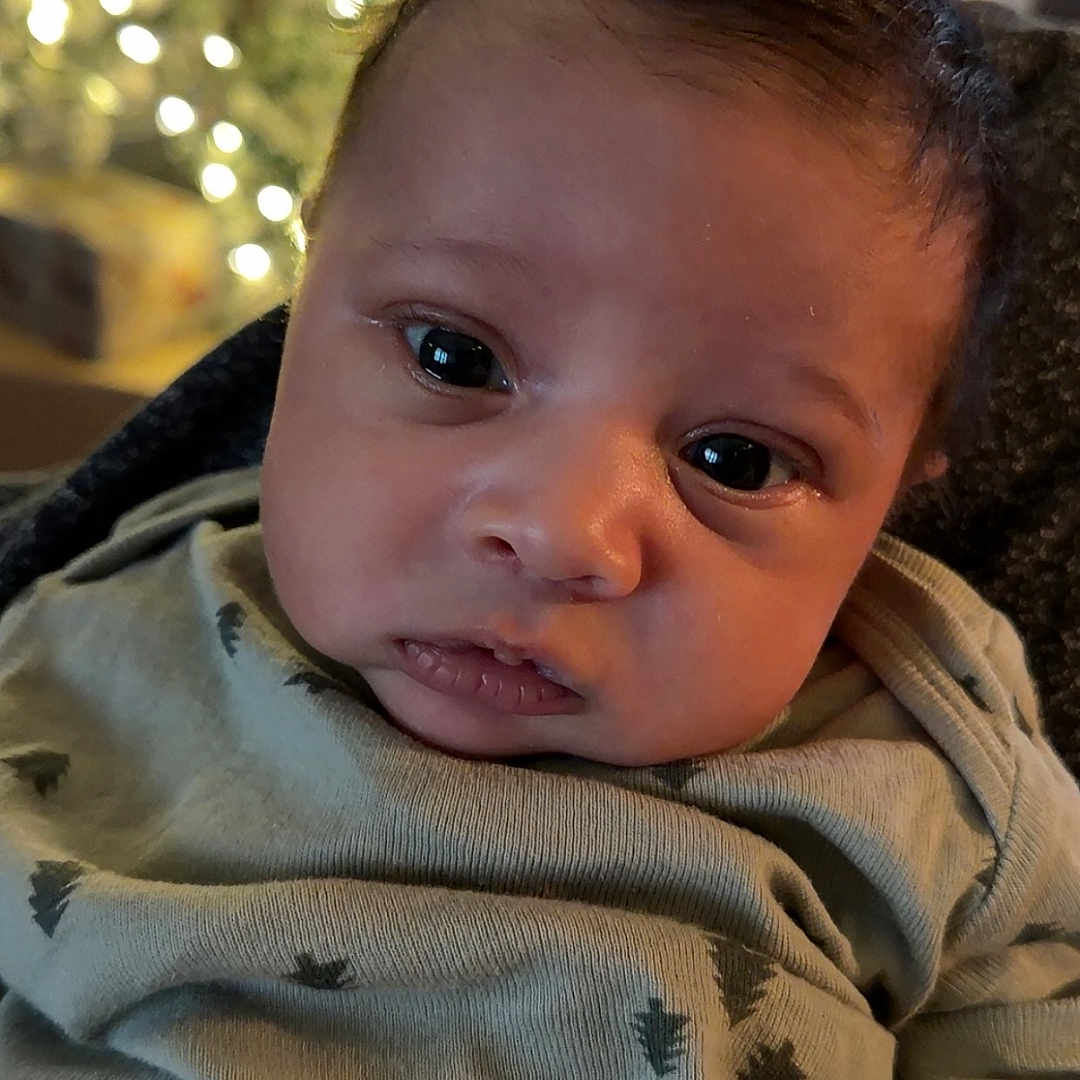 Malakai is registered to the contest to win money with this photo: baby, child, close_up, clothing, cozy, curious, cute, expression, eyes, fabric, face, holiday_lights, indoors, infant, newborn, pattern, portrait, soft_lighting, tree_print, warm