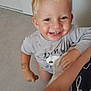 child, toddler, blonde_hair, smiling, indoor, floor, gray_floor, sandal, hand, arm, shirt, tattoo, person, happy, looking_up, short_sleeves, casual_clothing, skin, face, standing