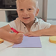 Naël a rejoint le concours — aidez-le/la à gagner de superbes lots ! child, boy, smiling, drawing, pen, paper, table, kitchen, bread, blonde_hair, white_shirt, appliance, microwave, shelf, container, indoor, food, happy, casual_clothing, person