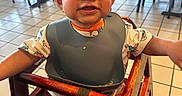 Avian is registered to the contest to win money with this photo: baby_shoes, bib, casual, child, clothing, cute, face, happy, high_chair, indoor, person, restaurant, seatbelt, short_hair, smiling, table, tile_floor, toddler, wooden_chair, young_child