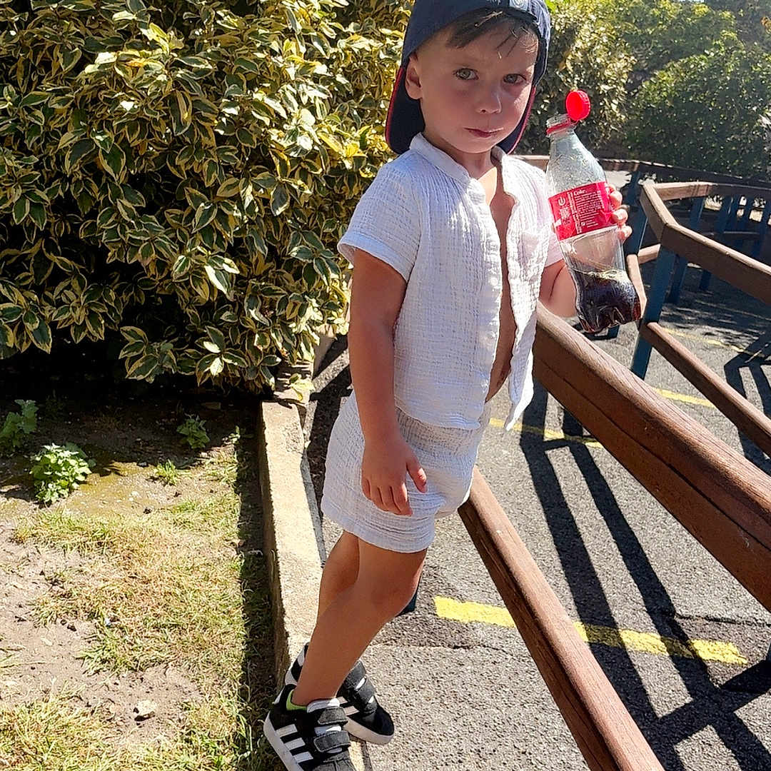 Leevay participe au concours pour gagner de l'argent avec cette photo : child, boy, outdoor, sunlight, greenery, soda_bottle, cap, white_clothing, black_sneakers, curb, sidewalk, wooden_railings, shadow, summer, casual, young, standing, nature, playful, daytime