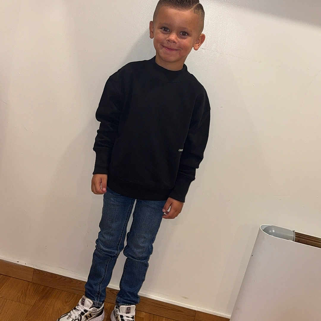 Leeroy a rejoint le concours — aidez-le/la à gagner de superbes lots ! child, boy, standing, casual_clothing, black_sweatshirt, blue_jeans, sneakers, indoor, wooden_floor, white_wall, heater, smiling, short_hair, fashion, portrait, person, happy, modern, youth, full_body