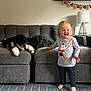 Elsie joined the competition — help win amazing prizes! toddler, child, dog, couch, living_room, rug, toy, smiling, barefoot, lamp, picture_frame, gray_couch, happy, indoor, home, furniture, blond_hair, carpet, decor, pet