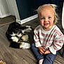 Elsie is registered to the contest to win money with this photo: toddler, child, dog, indoor, wooden_floor, striped_sweater, jeans, pigtails, smiling, pet, flooring, home, cute, sitting, casual_clothing, light_hair, blue_eyes, relaxed, domestic_animal, companion