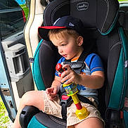 Axel is registered to the contest to win money with this photo: toddler, child, car_seat, toy, baseball_cap, blue_shirt, shorts, vehicle_interior, open_car_door, seatbelt, greenery, window, daylight, curious, sitting, playing, person, indoors, safety, casual_clothing