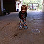 child, barn, toy_wagon, car, gate, concrete_floor, sunflower_pants, long_sleeve_shirt, smiling, indoor, daylight, rustic, wooden_walls, playful, person, young_child, brown_hair, casual_clothing, open_door, vehicle