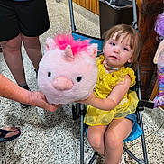 Oakley is registered to the contest to win money with this photo: toddler, child, stroller, yellow_dress, stuffed_unicorn, toy, indoor, floor, wood_paneling, person, hand, footwear, shorts, necklace, pink, blue, casual_clothing, seated, playful, expression